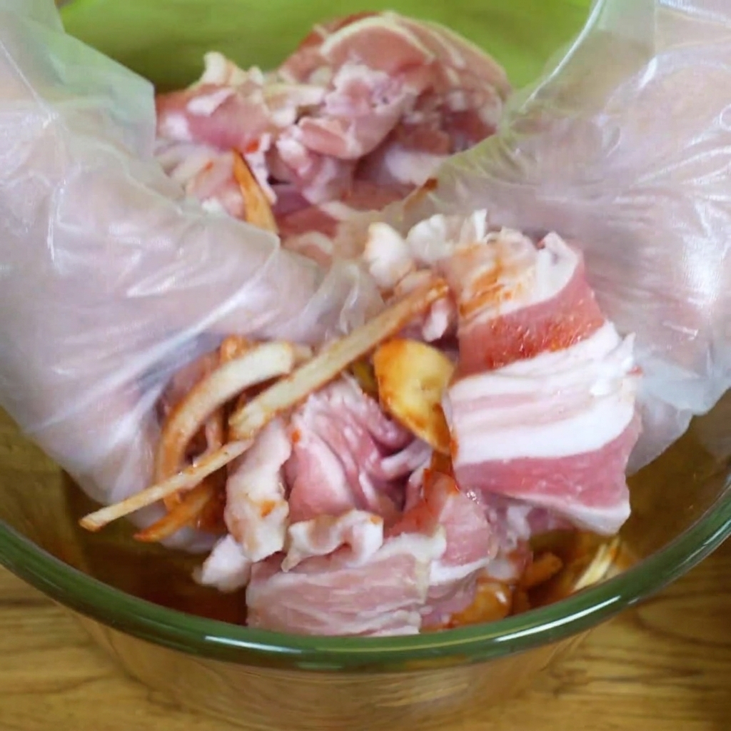 Hands wearing plastic gloves mixing sliced pork belly with marinade and vegetables in a glass bowl.