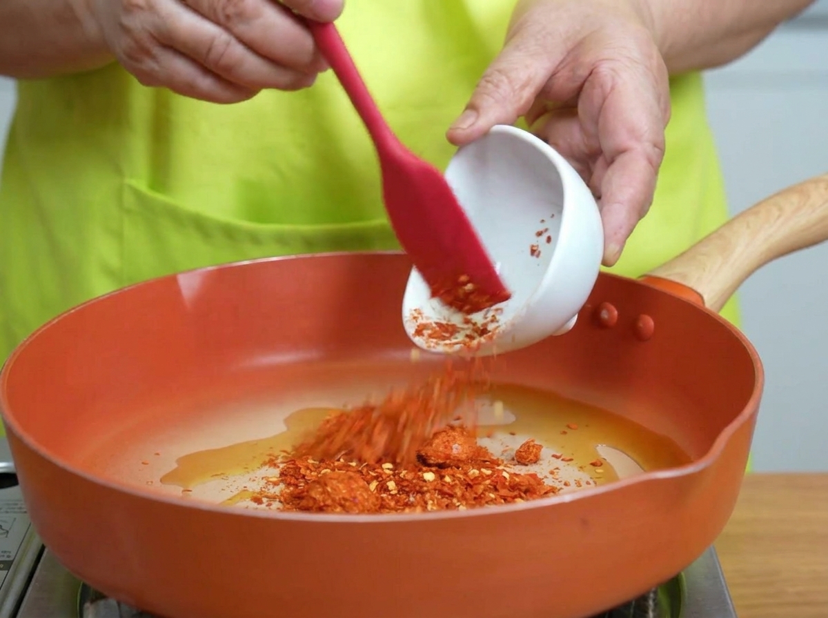 Chili flakes being stirred into shimmering hot oil in a pan.