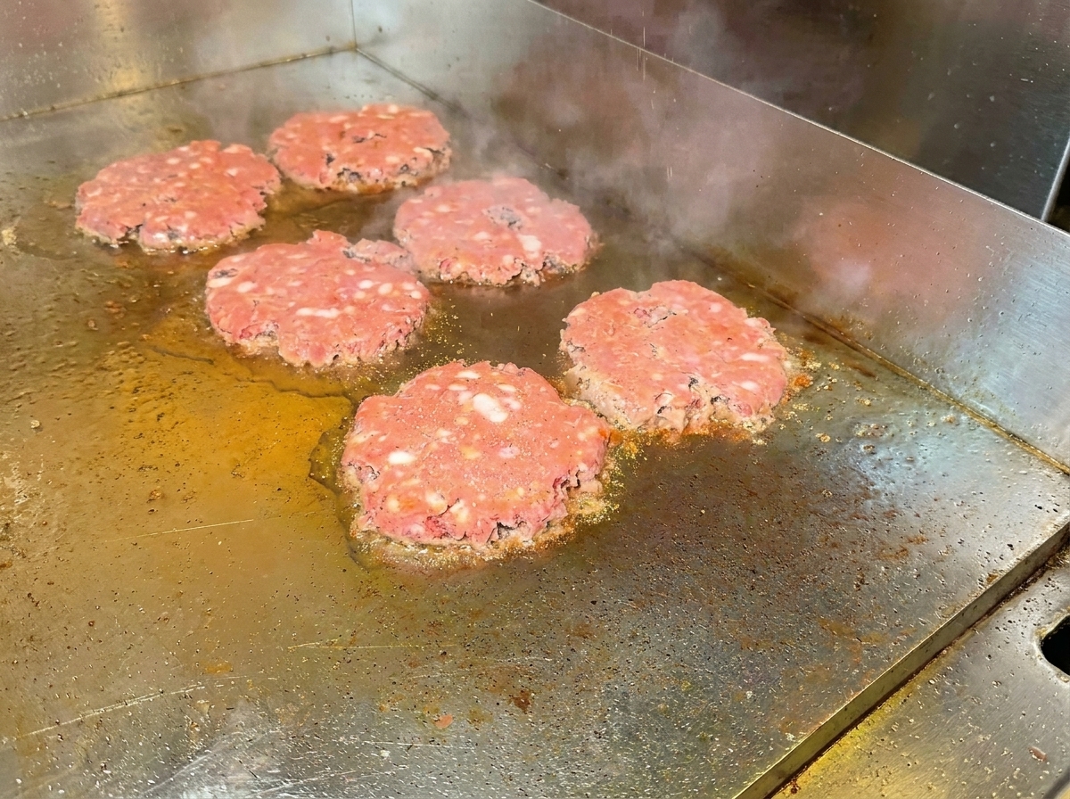 Beef patties being flipped on the griddle, showing a browned surface and seasoning.