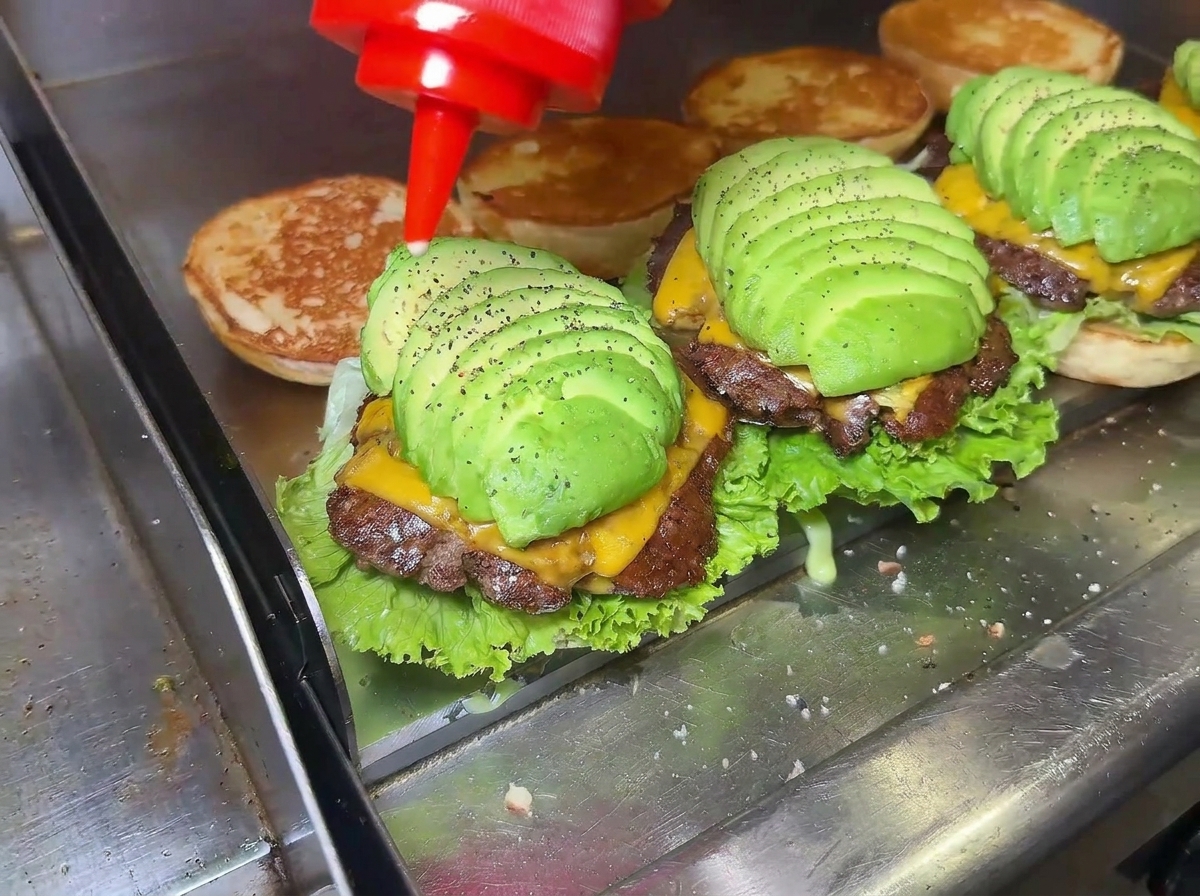 A close-up of a hand squeezing a red sauce bottle, drizzling sauce onto a perfectly layered avocado cheeseburger on a griddle.