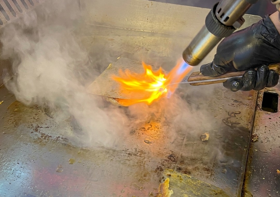 A beef patty on the griddle being charred with a kitchen blowtorch, emitting steam and sparks.