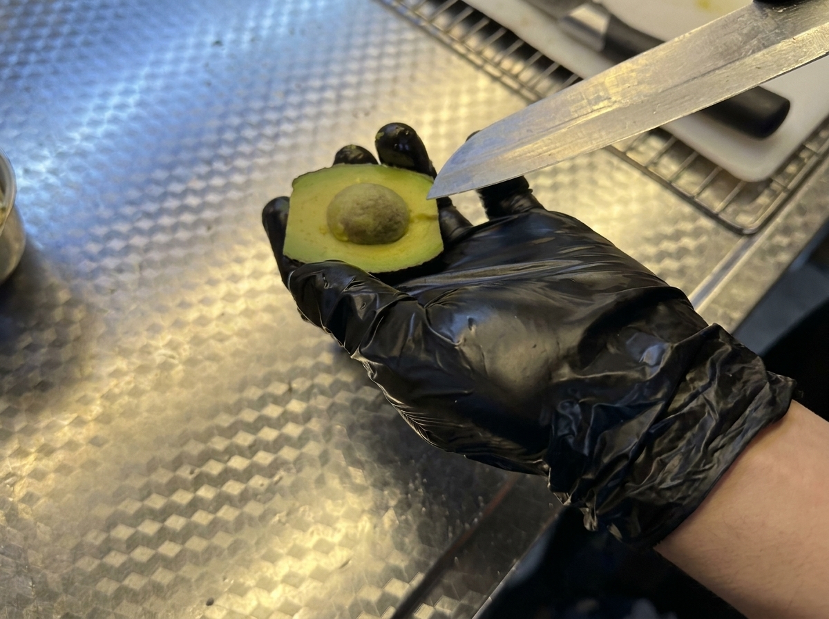A hand wearing a black glove holding an avocado half while a knife is used to remove the pit.