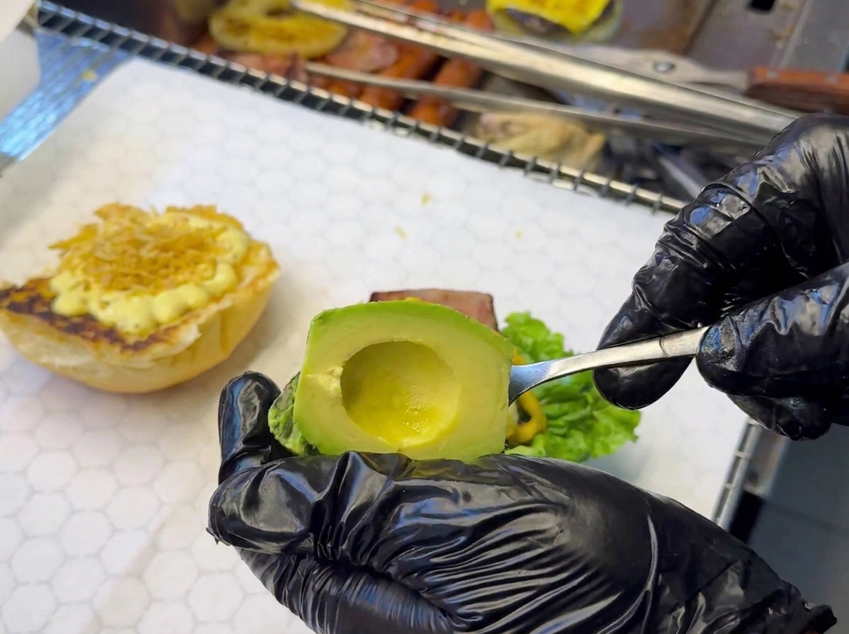 A hand in a black glove using a spoon to place a slice of avocado onto a burger base that already includes lettuce and sauce.