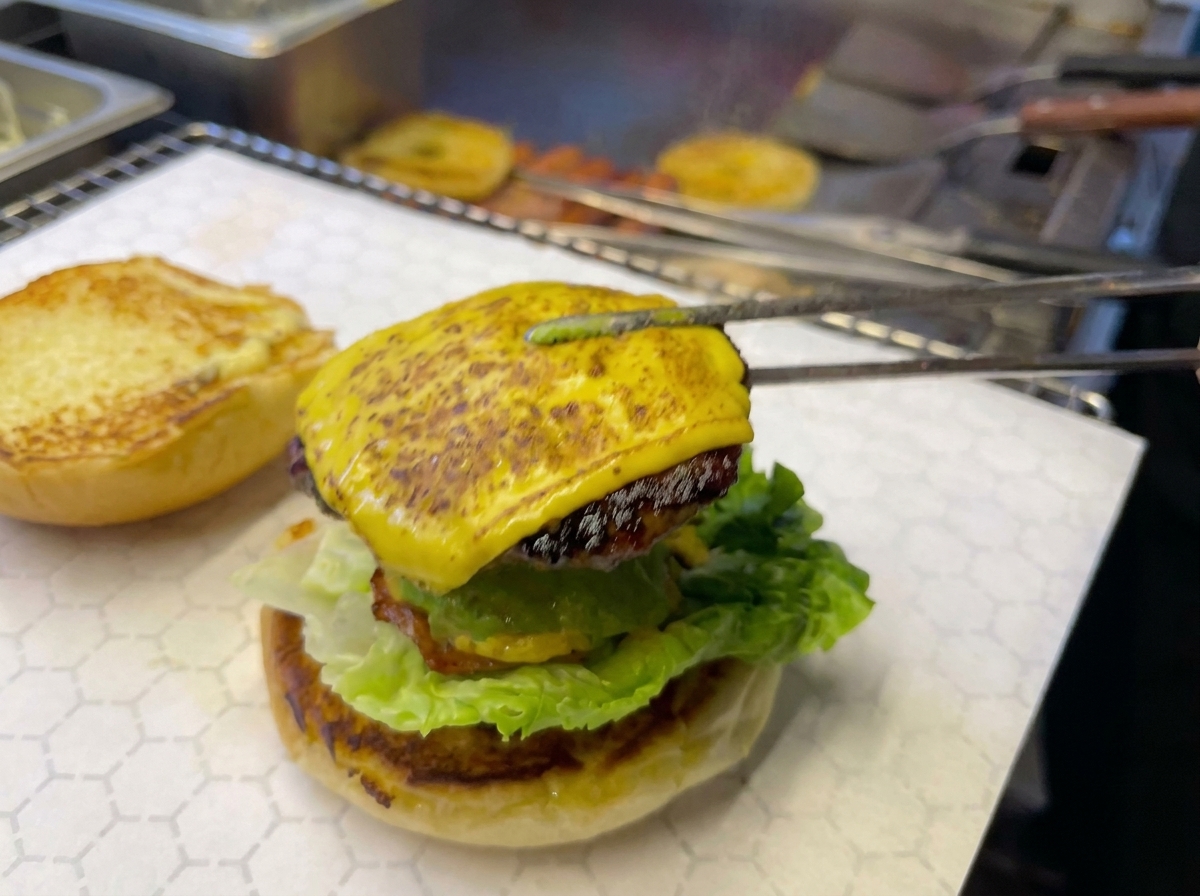 Kitchen tongs placing a cheesy beef patty topped with a pineapple ring onto a burger that contains avocado and lettuce.