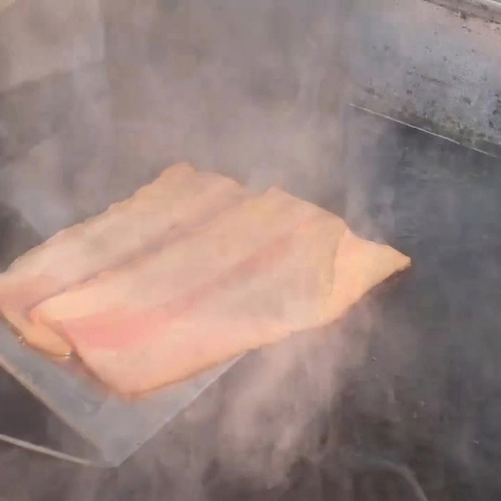 Strips of bacon frying on a hot griddle, emitting steam.