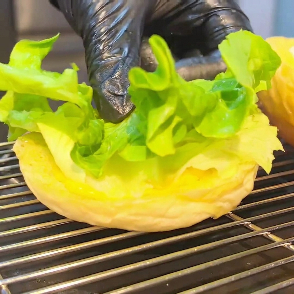 A half-assembled burger on the bottom bun, featuring lettuce, tomato, a cheesy beef patty, and bacon strips.