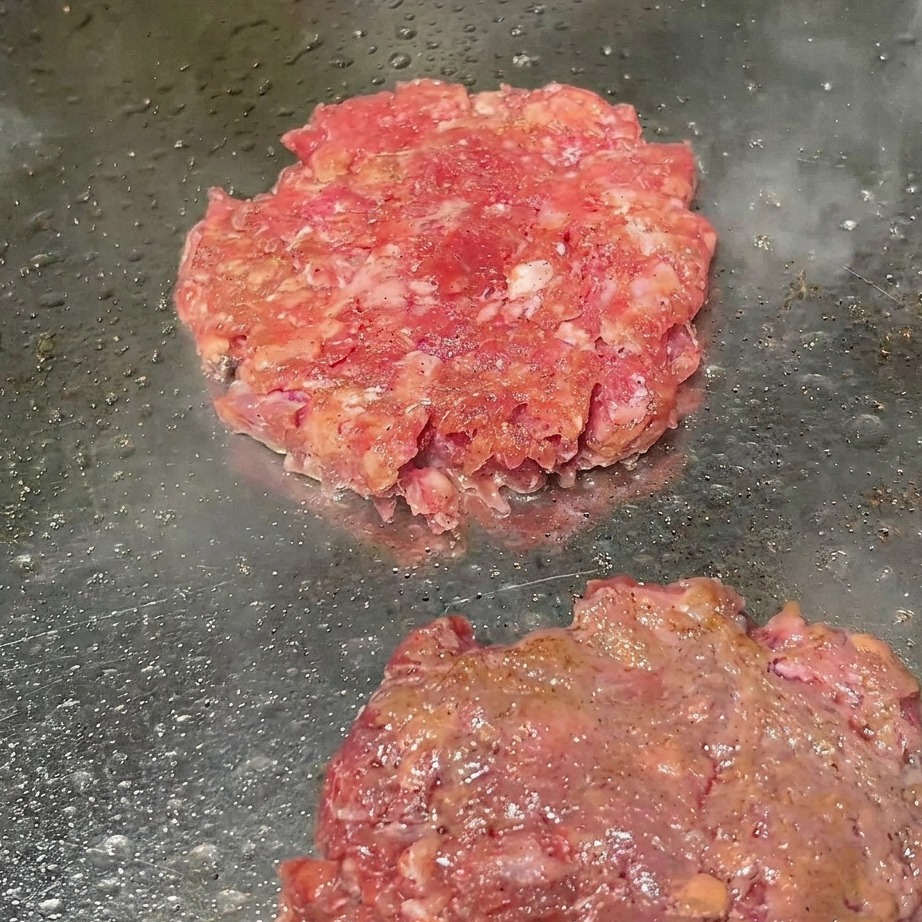 Beef patties on a grill being dusted with freshly ground black pepper.