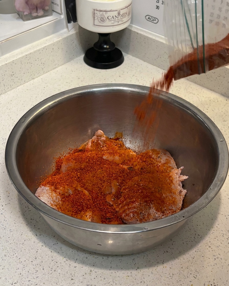 Raw chicken thighs being seasoned with spice powder and milk in a large metal bowl.