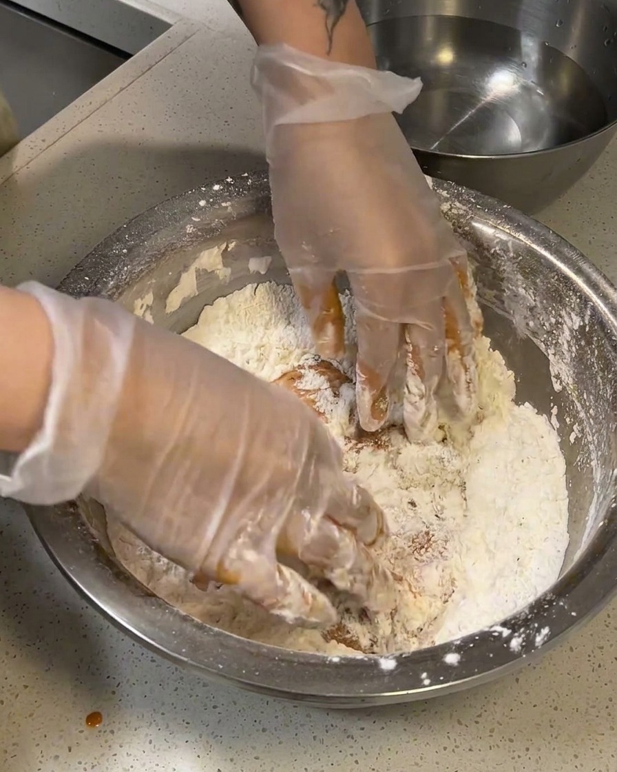 Raw marinated chicken being firmly pressed into a bowl of dry flour breading.