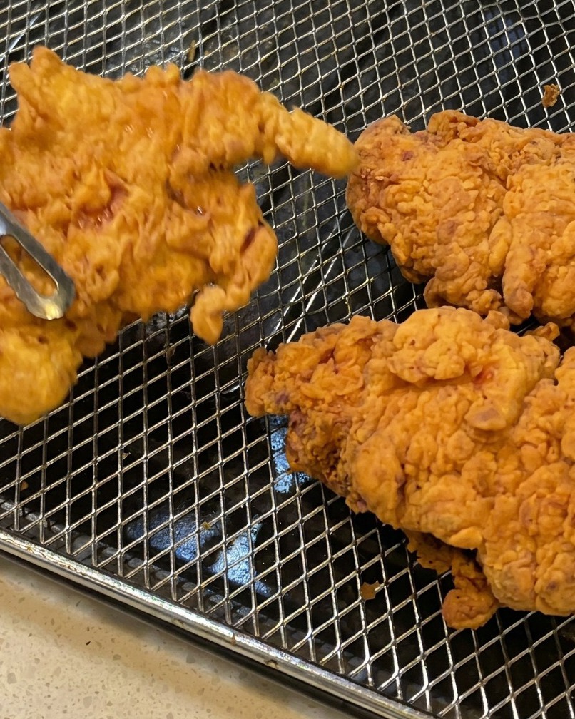 Fried chicken thighs being lifted out of the oil with metal tongs, showing crispy, flaky breading.