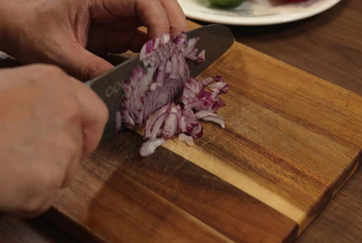 Red onion being finely diced on a wooden cutting board with a knife.