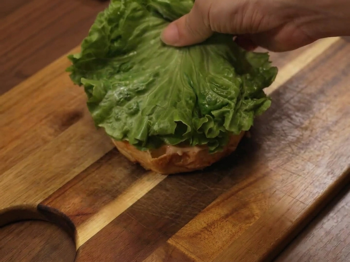 A lettuce leaf being placed on the bottom bun during the assembly of the burger.