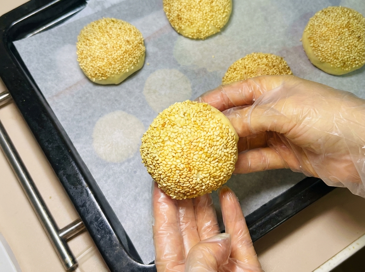 A gloved hand arranging a sesame-coated dough ball on a parchment-lined baking tray alongside other coated buns.