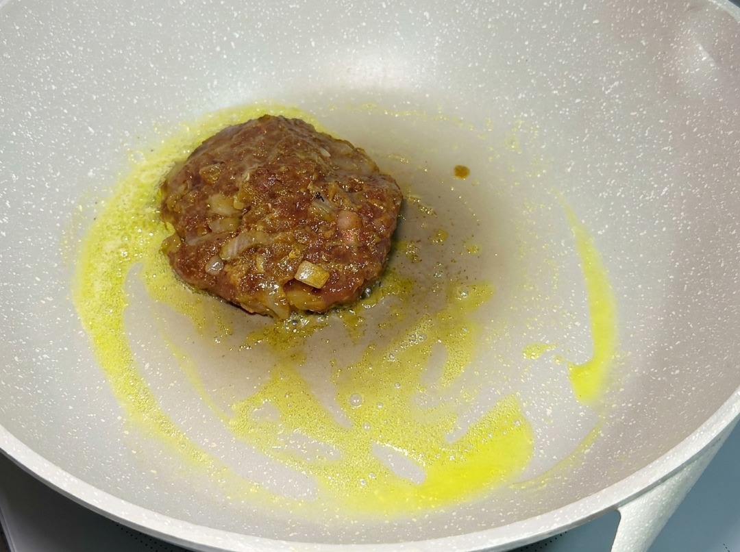 A raw seasoned beef patty frying in a light-colored pan over low heat, surrounded by melted fat.