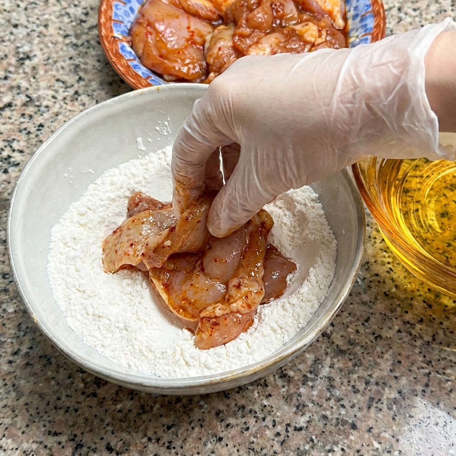 A marinated raw chicken thigh being pressed into a bowl of dry flour mixture.
