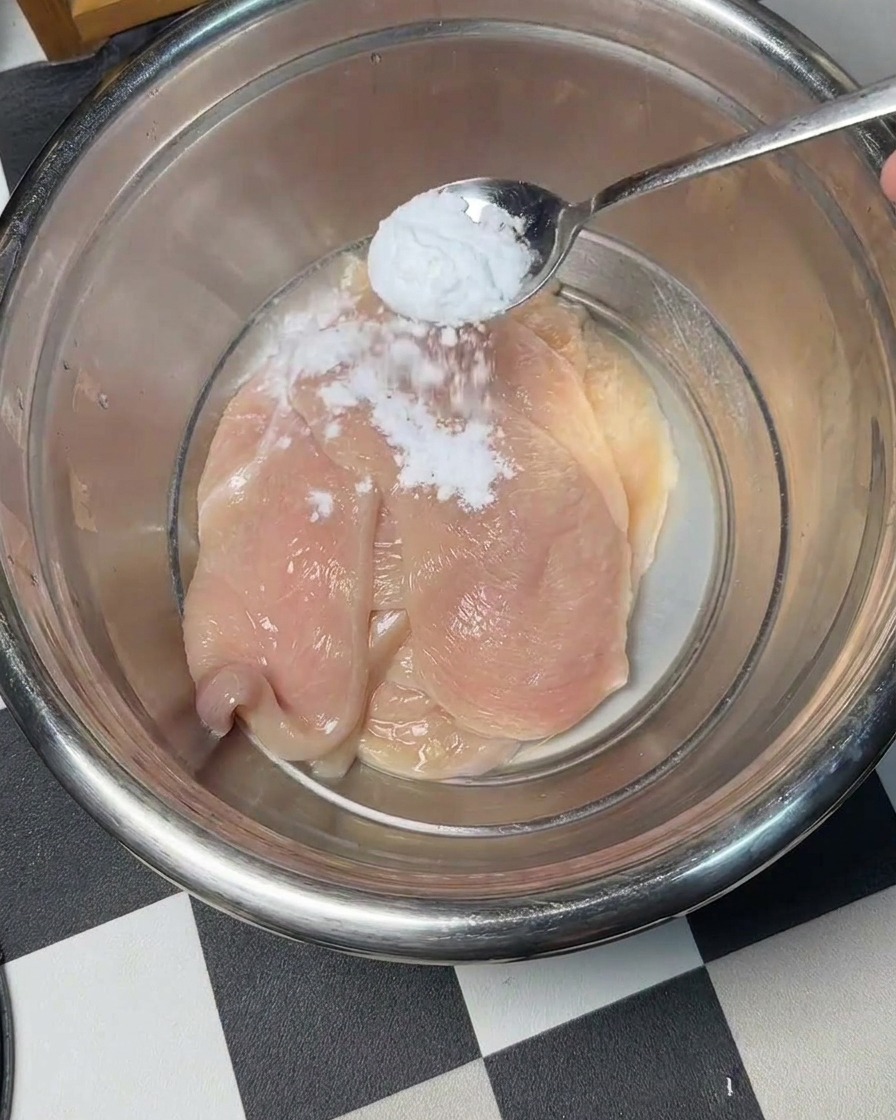 Baking soda being sprinkled over raw chicken slices in a mixing bowl.