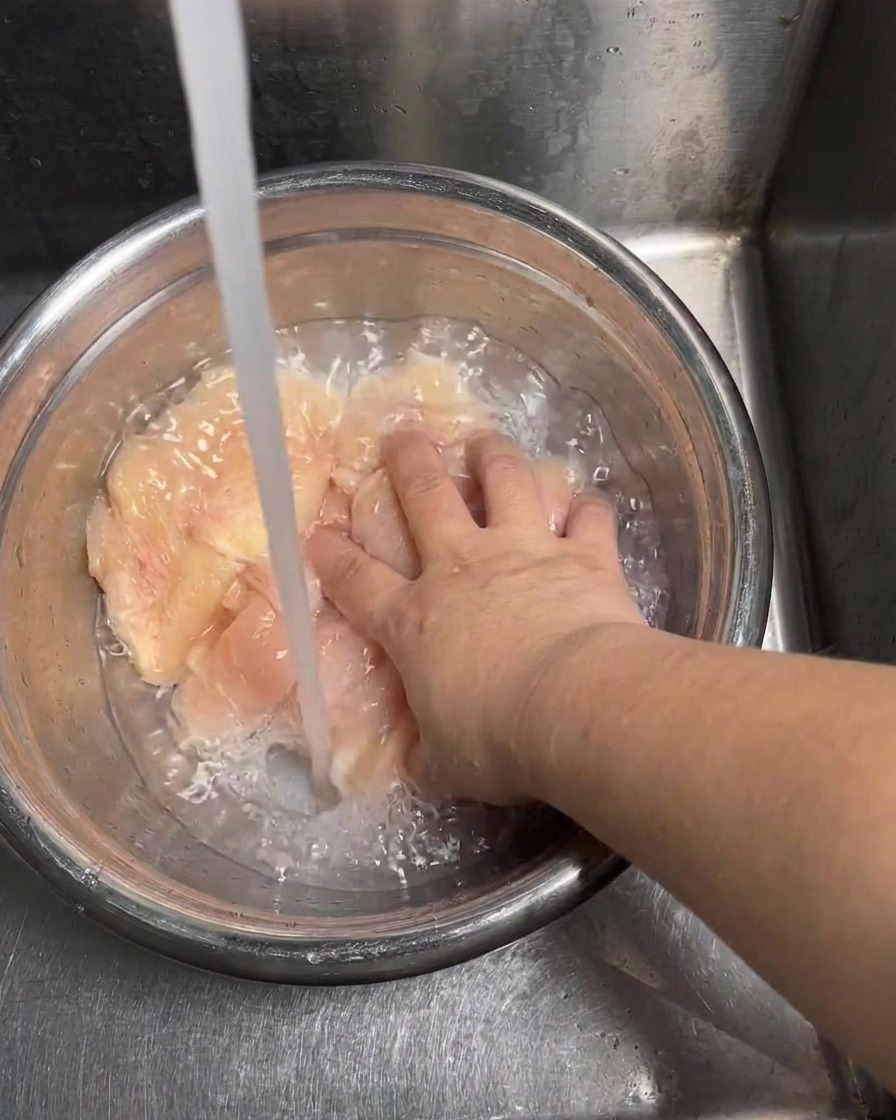 Raw chicken slices being rinsed under a steady stream of cold water.