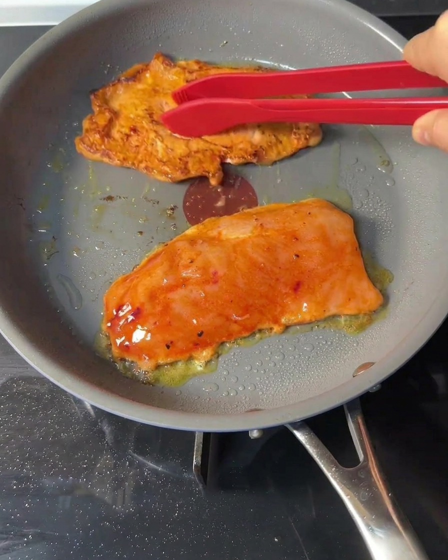 Red silicone tongs flipping browning chicken patties inside a frying pan.