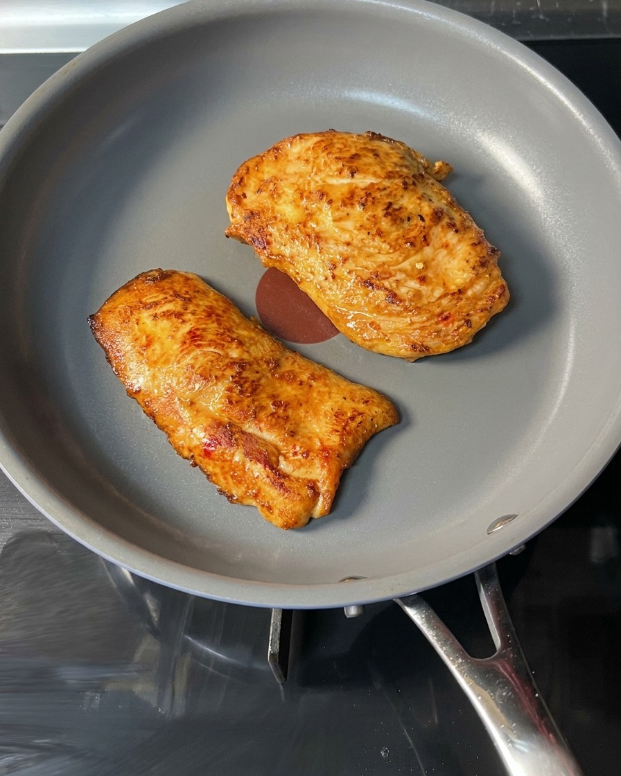 A cooked, golden-brown chicken patty being lifted from a pan with red tongs.
