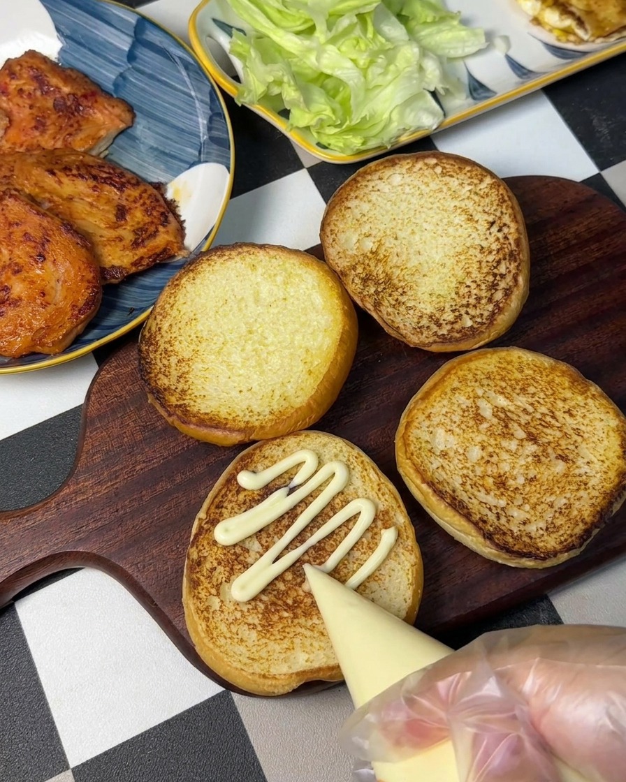 A hand using a piping bag to squeeze mayonnaise in a zigzag pattern onto a toasted burger bun.
