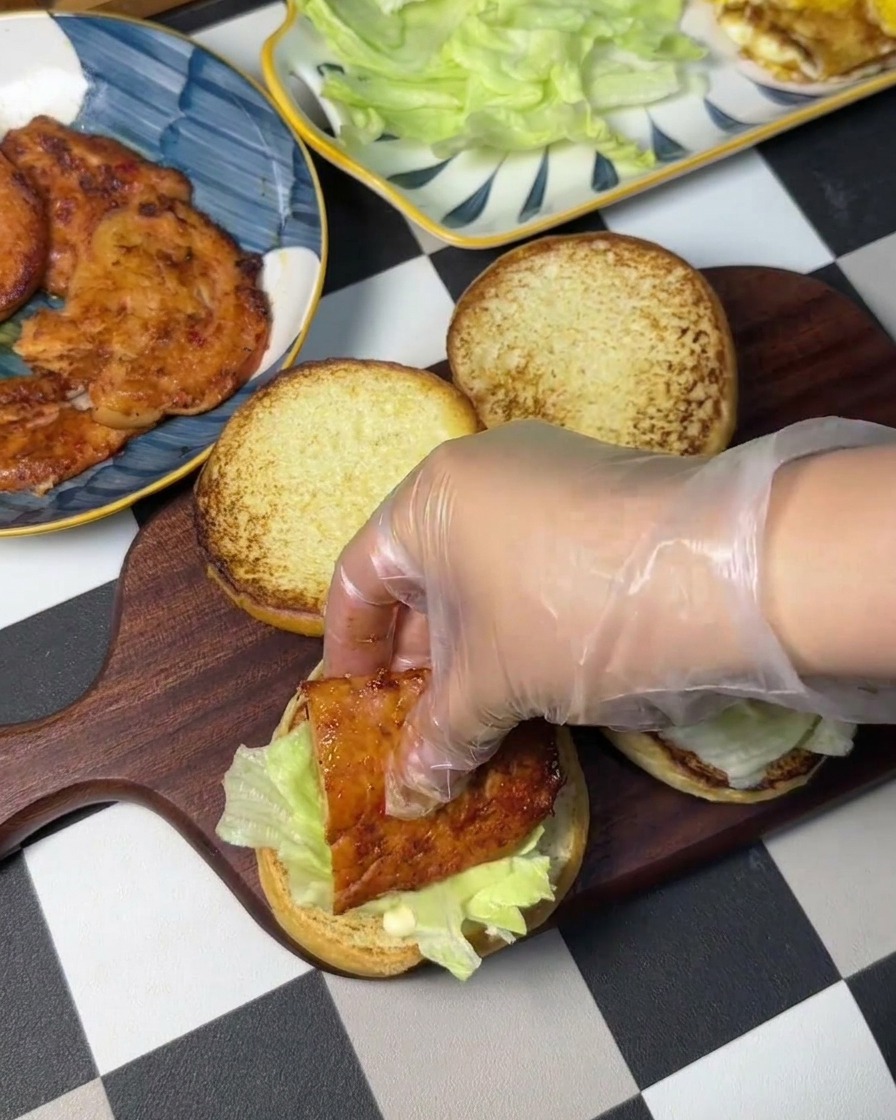 A gloved hand placing a pan-fried chicken patty over a bed of fresh lettuce on a burger bun.