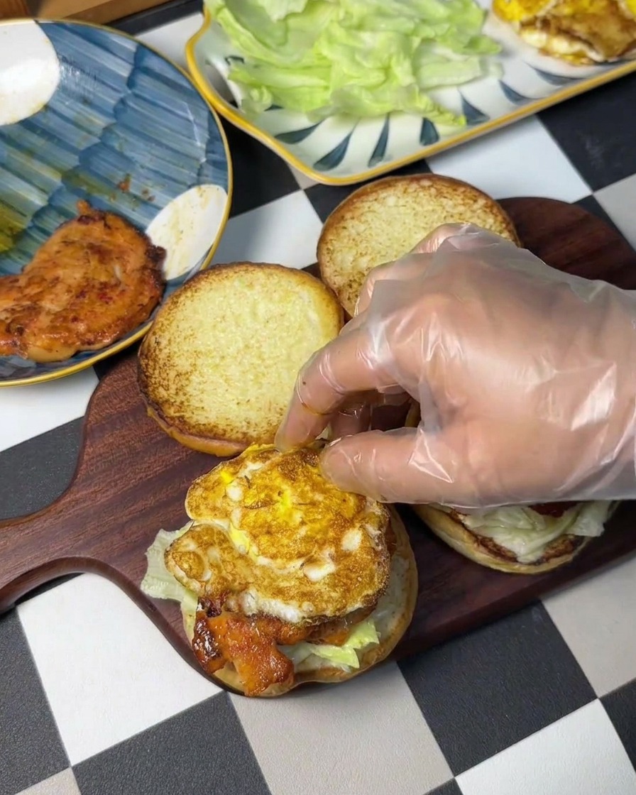 A gloved hand placing a pan-fried egg with crispy edges onto a chicken patty.