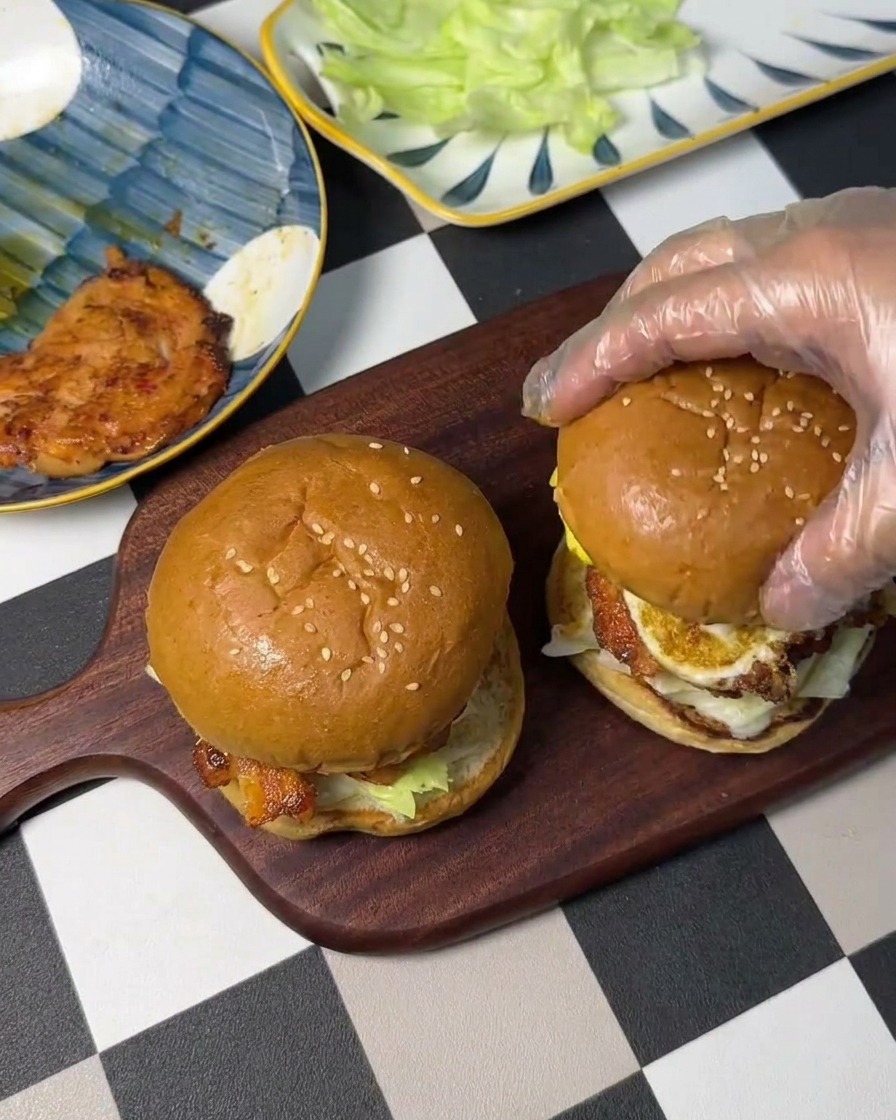 A gloved hand placing a toasted top burger bun over a stack of egg, chicken, and lettuce to complete a sandwich.