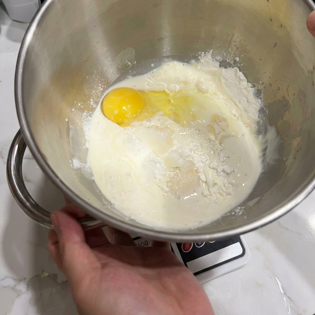 A stand mixer bowl containing flour, a cracked egg, and milk, ready to be mixed.