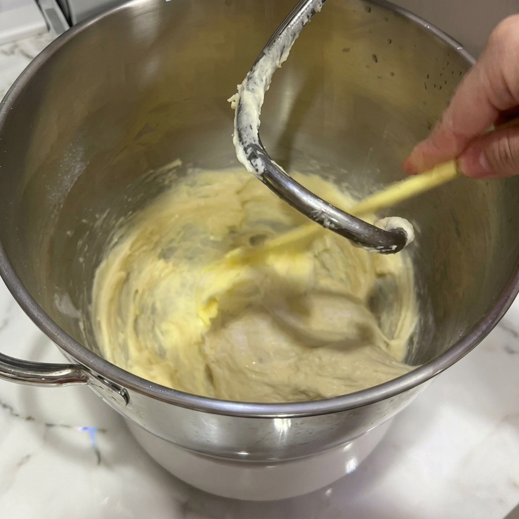A block of softened yellow butter being added to a partially formed dough inside a stand mixer.