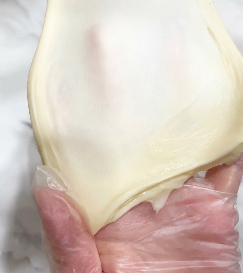 A piece of smooth, elastic dough being stretched thin by hand to show light passing through.