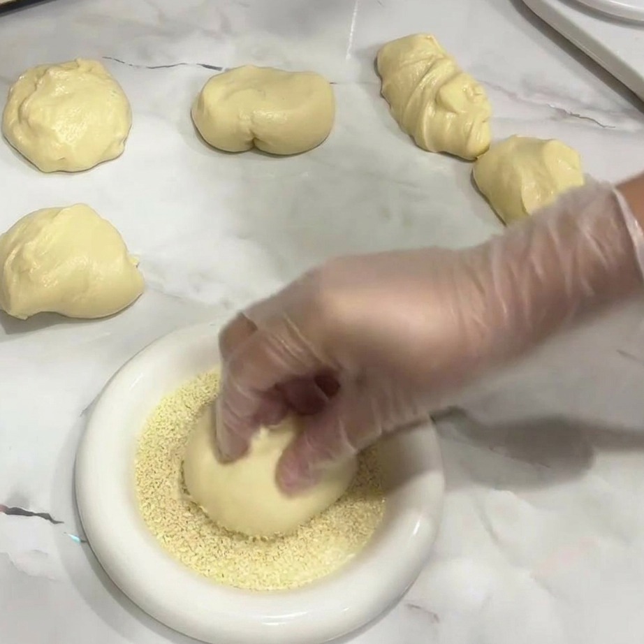 A hand placing a smooth, round dough ball into a plate of white sesame seeds.