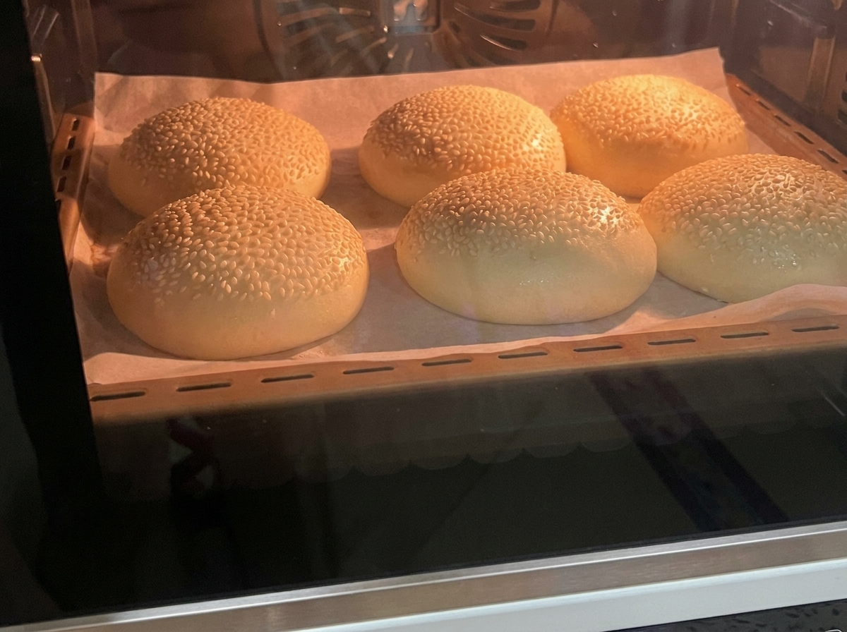 Six freshly baked, golden-brown sesame hamburger buns sitting on a parchment-lined tray inside an oven.