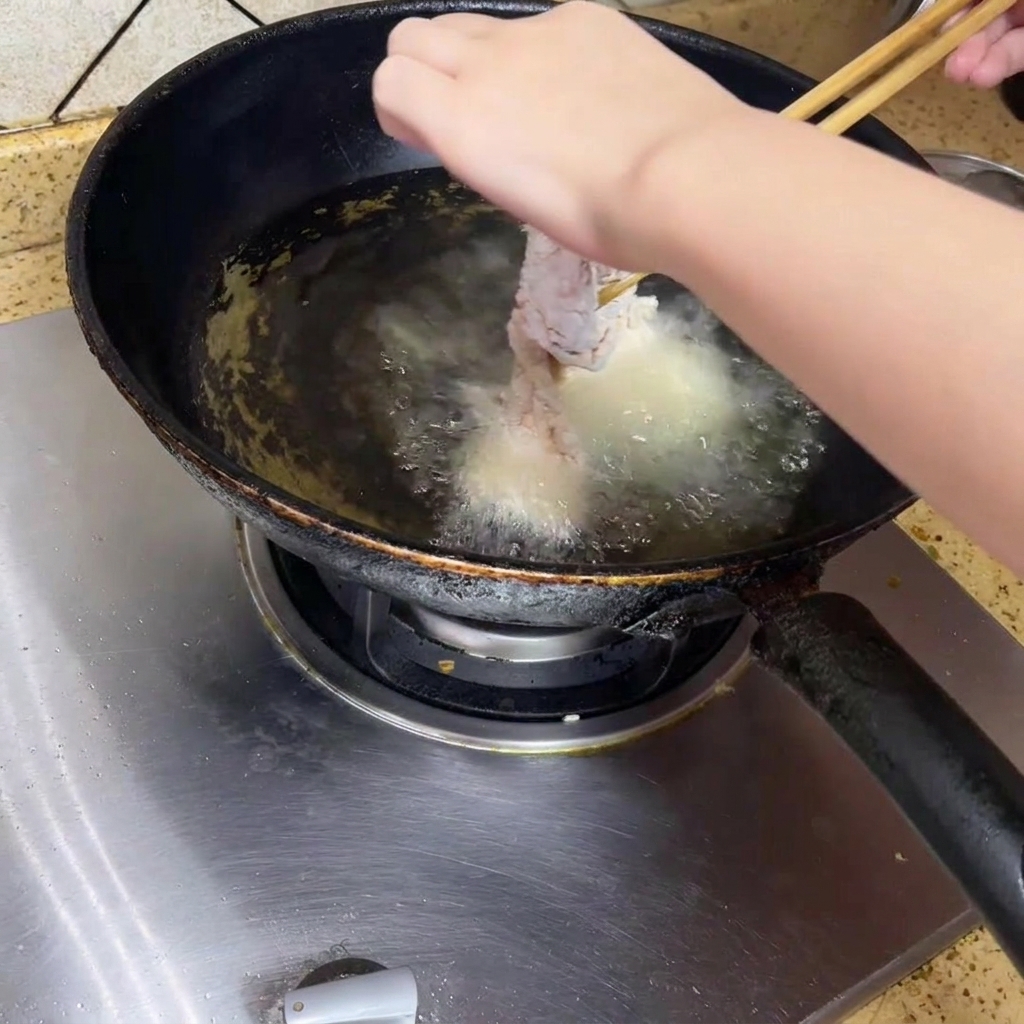 A person using chopsticks to gently lower a piece of breaded chicken into a black wok filled with hot, bubbling oil.