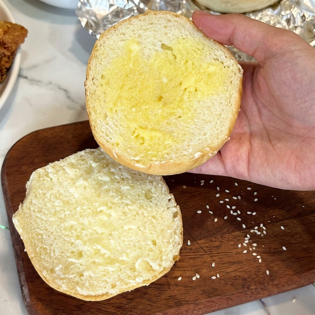 The cut half of a sesame hamburger bun coated in melted butter, held over a wooden board.
