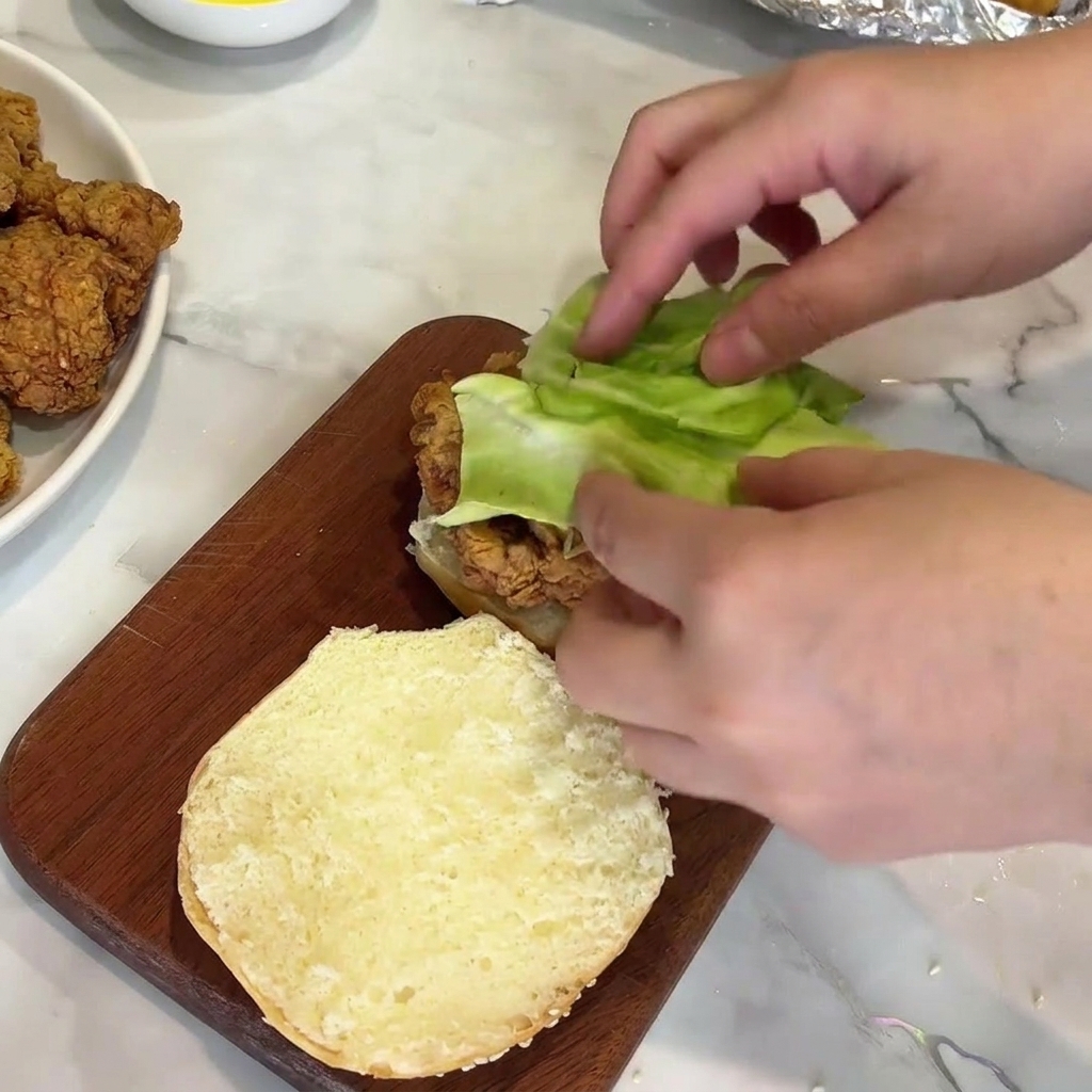 Hands placing a piece of fresh green lettuce on top of a crispy fried chicken patty on a bun.