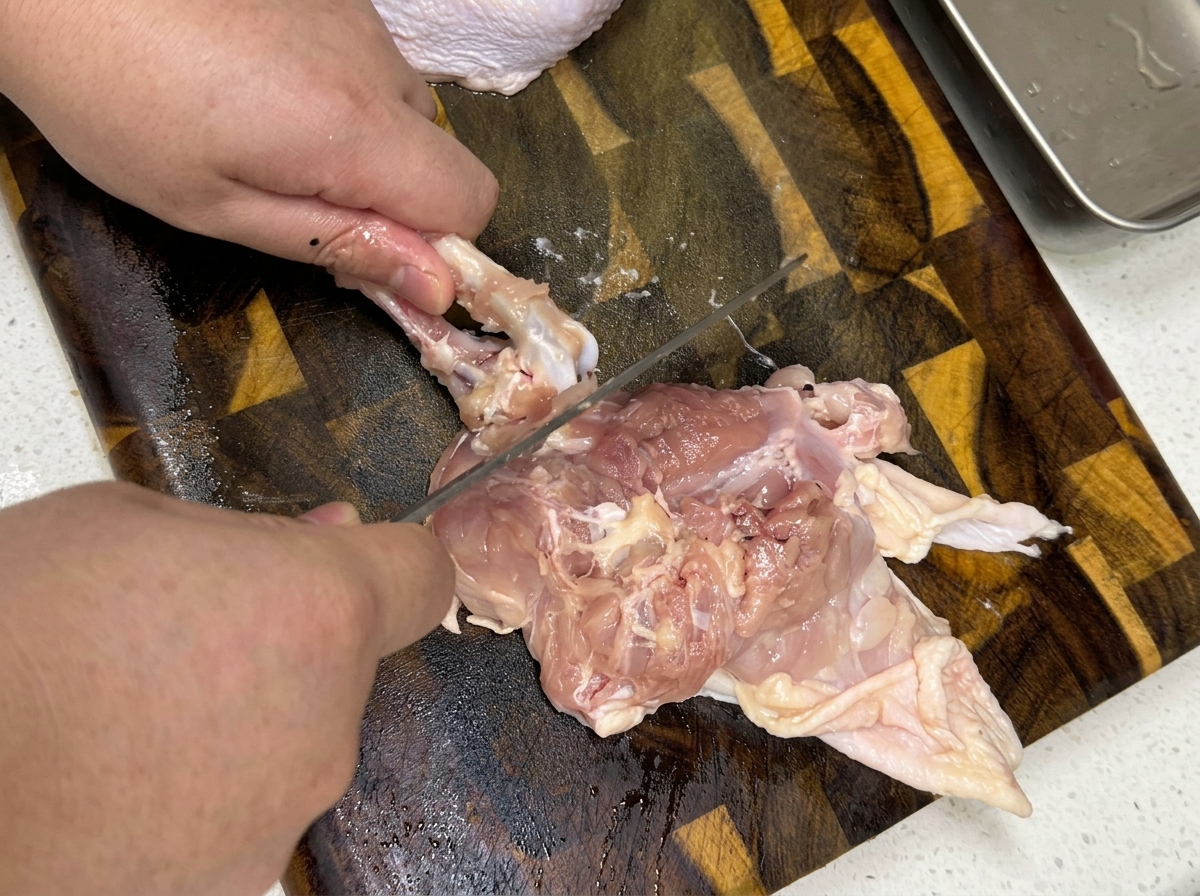 A close-up of hands deboning a raw chicken thigh on a wooden cutting board.