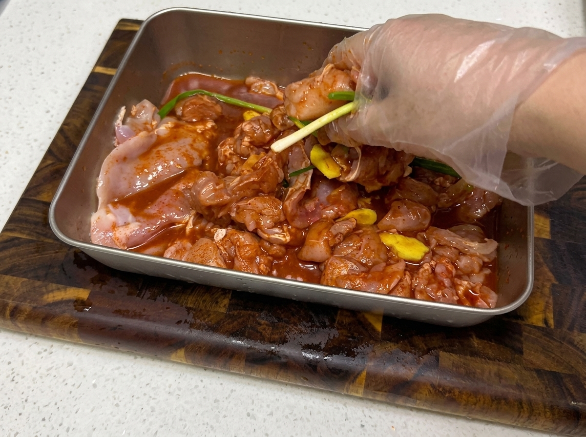 Hands mixing marinade into the diced chicken in a metal tray.