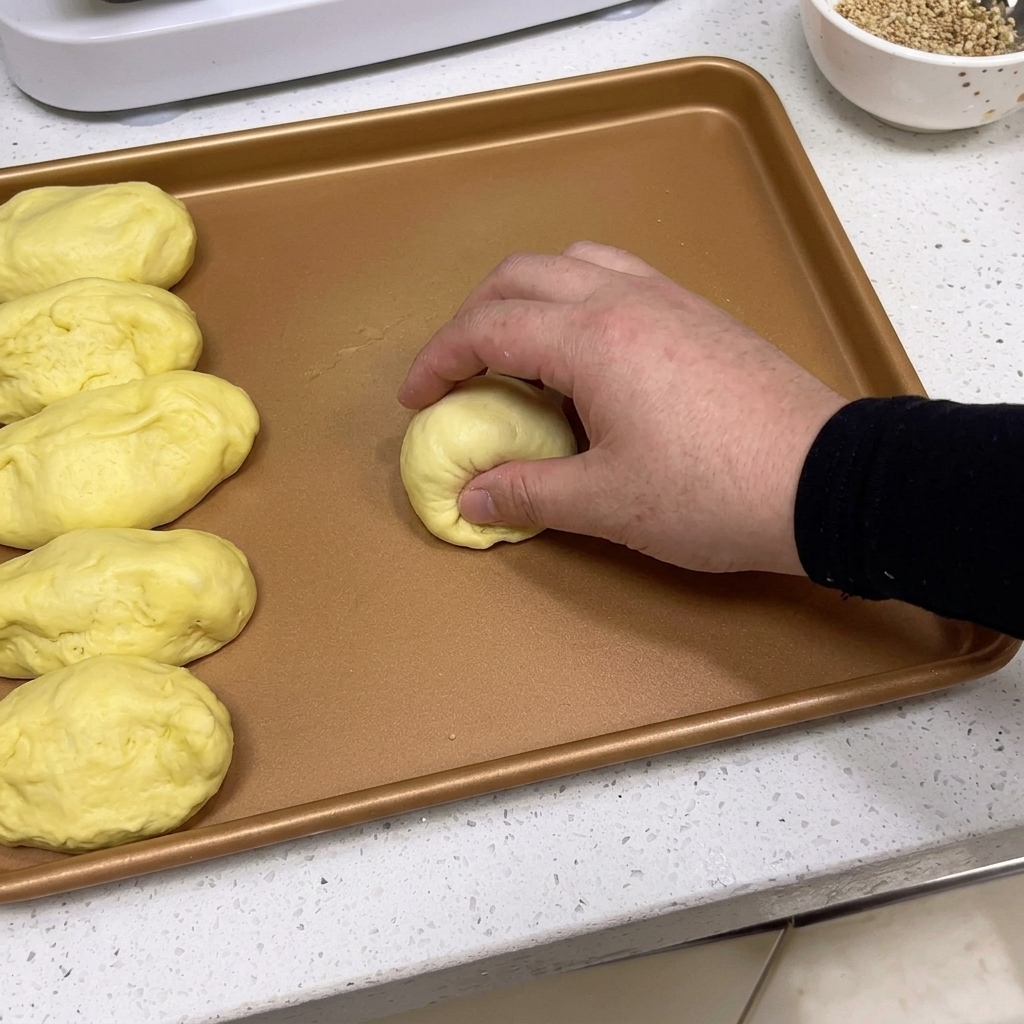 Hands shaping a portion of dough into a smooth, round ball on a tray.