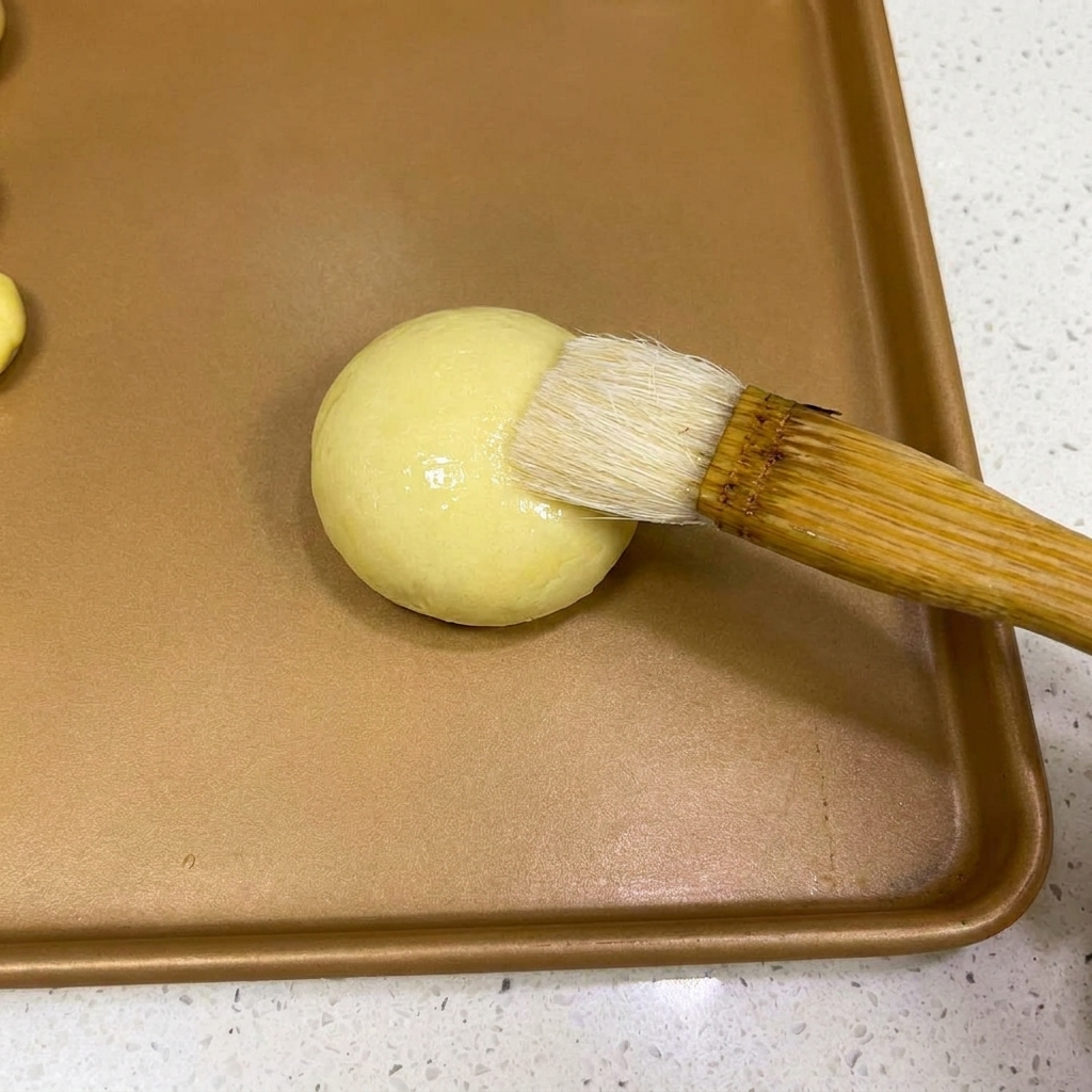 A pastry brush applying a light layer of water to the top of a rounded dough ball.
