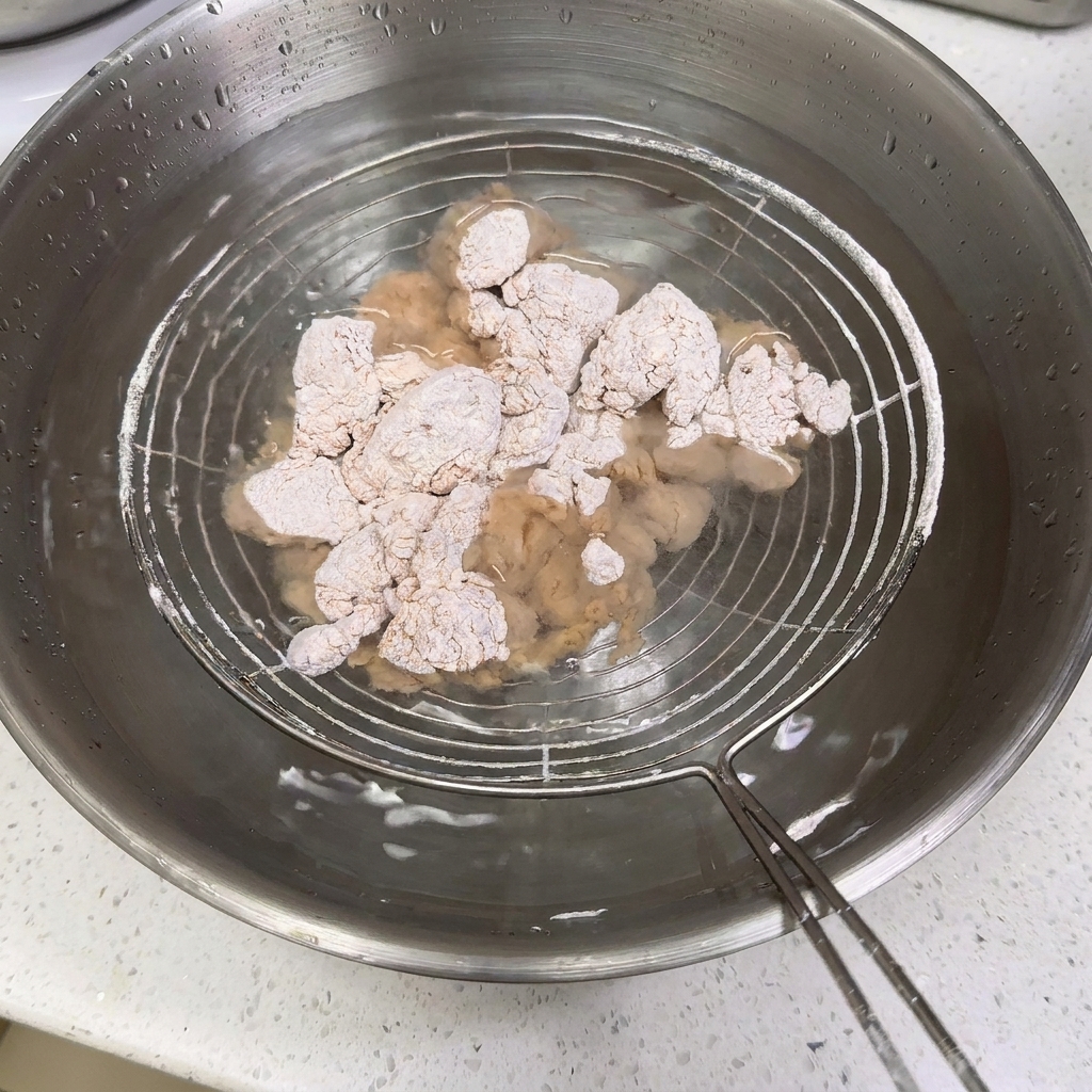 Flour-coated chicken pieces submerged in a stainless steel bowl of water on a wire skimmer.