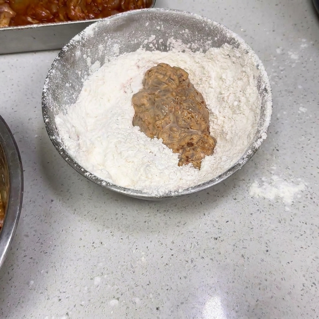 A piece of wet, battered chicken being tossed in a silver bowl filled with dry white flour.
