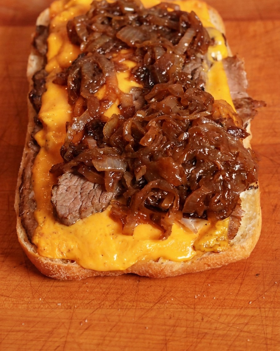 Rich, dark brown caramelized onions being spooned over yellow cheddar cheese on a sandwich base.