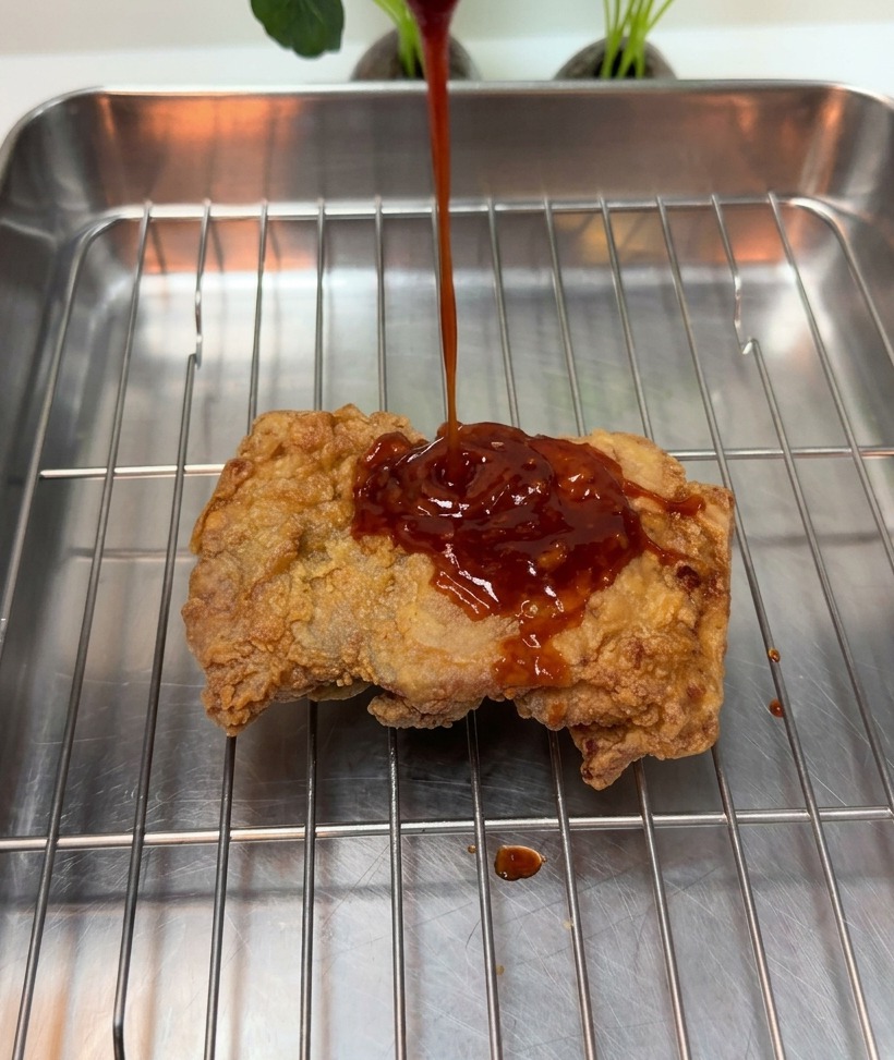 Thick red sauce being poured over a crispy fried chicken piece on a metal rack.
