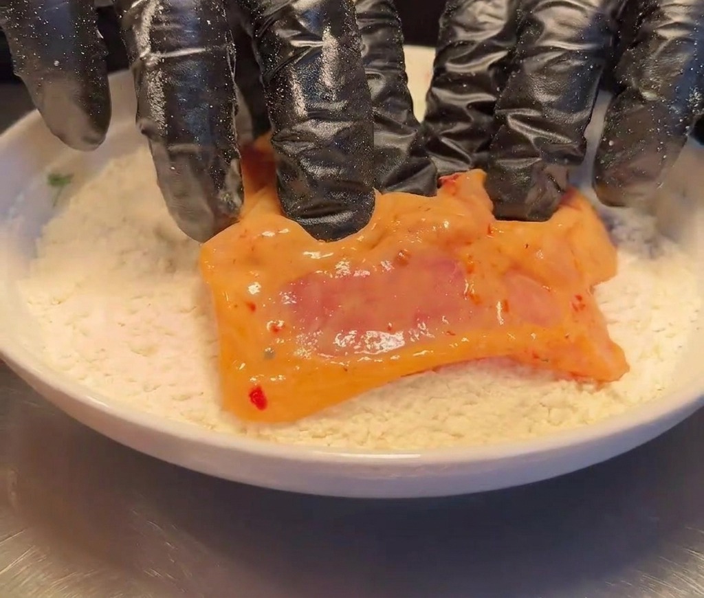 A raw, marinated chicken thigh being dredged in a plate of dry white flour.