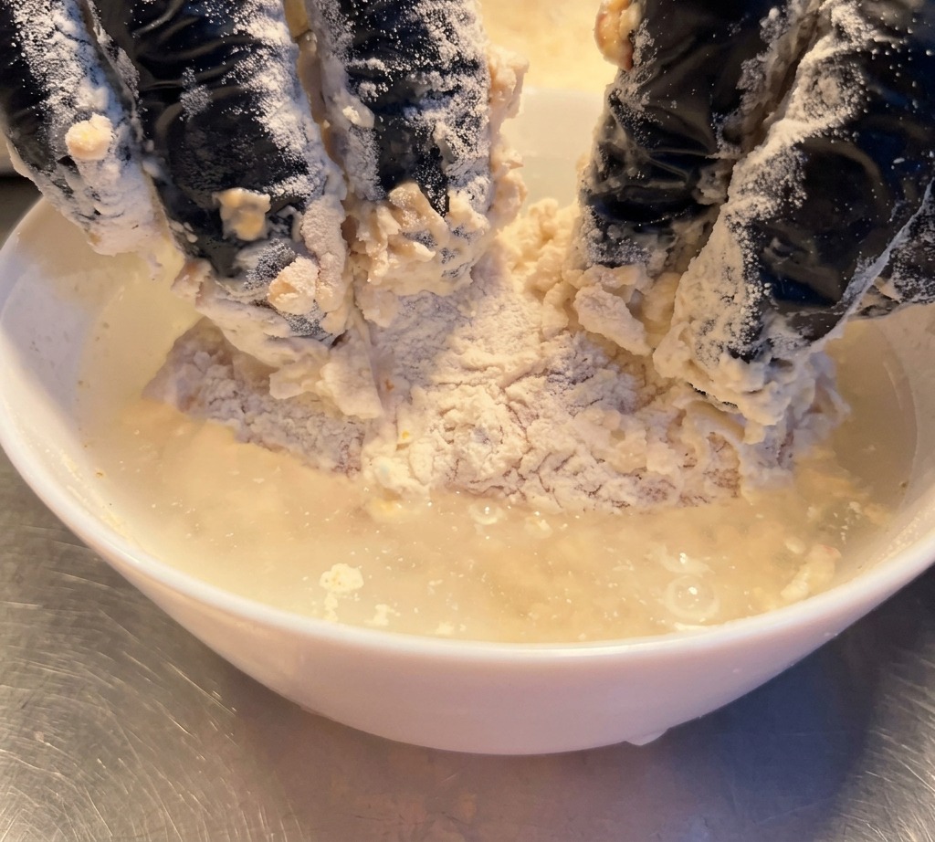 A flour-coated chicken piece being submerged briefly in a bowl of clear water.