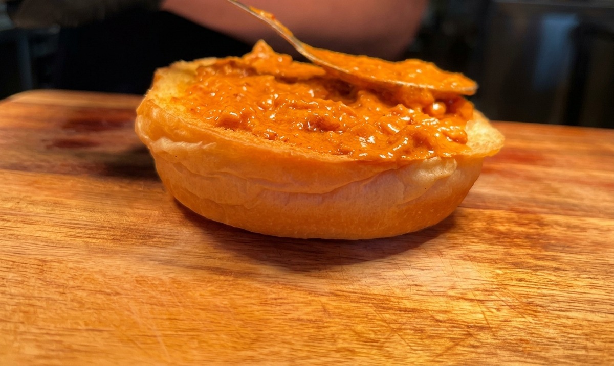 A spoon spreading orange spicy cream sauce onto a toasted bottom burger bun.