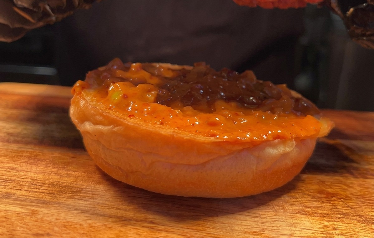 A gloved hand lowering a piece of crispy fried chicken onto a sauce-covered burger bun.