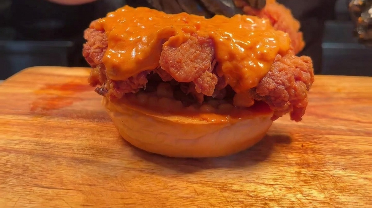 Gloved hands assembling a double chicken burger by stacking a second fried patty on top of the first.