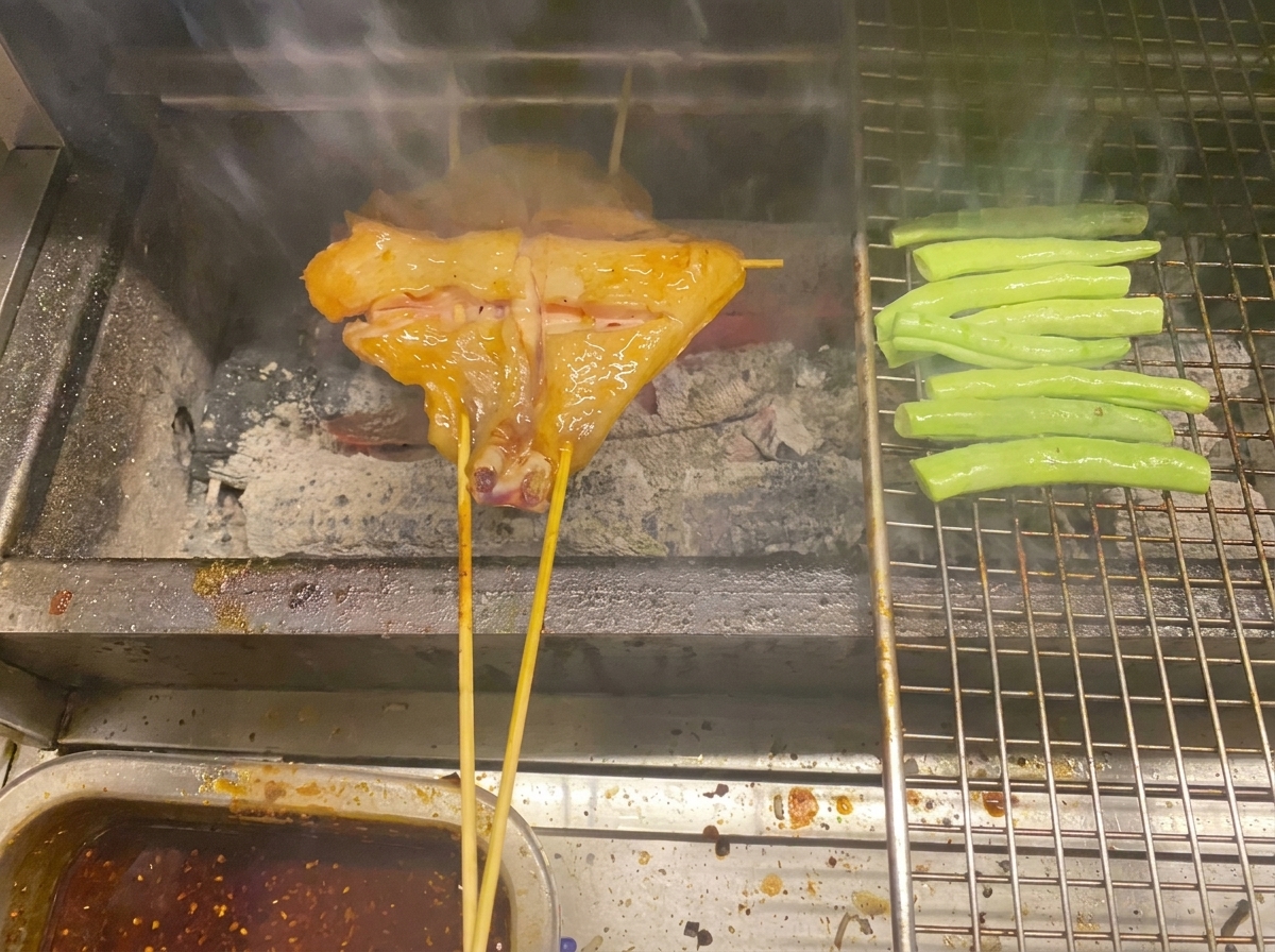 Skewered chicken and green beans cooking over charcoal on a metal grill.