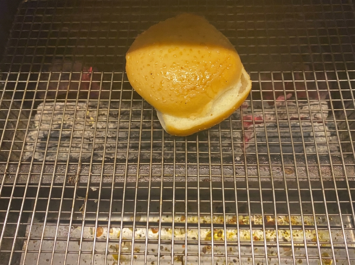 A burger bun half toasting face-down on a charcoal grill.
